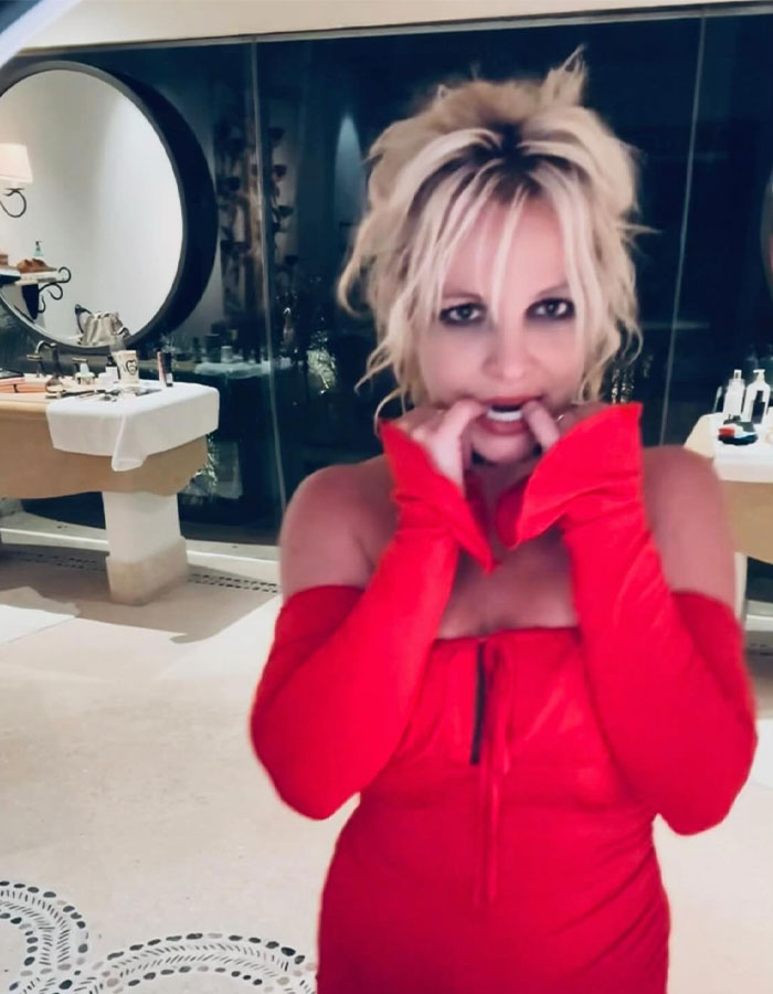 Woman in red dress with messy hair and chipped nails in a dimly lit room, looking toward the camera.